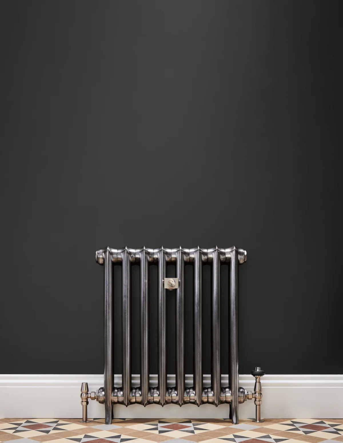 A wall-hung cast iron radiator in Full Polish on feet against a charcoal wall, with nickel valves, a white skirting board and geometric floor tiles.