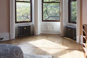 Two Mercury 4 Column cast iron radiators in a mid grey finish on feet beneath a sash bay window, with brass valves and a parquet floor.