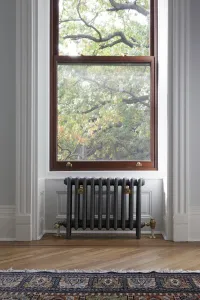 A plain Mercury 4 Column cast iron radiator in a grey finish on feet beneath a sash window, with brass valves and stays, on a herringbone floor.
