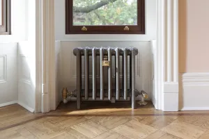 Plain Mercury 4 column cast iron radiator on feet in a metallic grey finish with brass valves, under a sash window on a parquet floor.