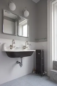 A narrow plain four-column Mercury cast iron radiator in a grey metallic finish on feet beside a wall-hung trough sink in a tiled bathroom.
