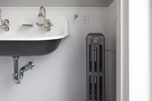 A plain four-column Mercury cast iron radiator in a mid grey finish with a wall stay, beside a wall-hung trough basin in a white-tiled bathroom.