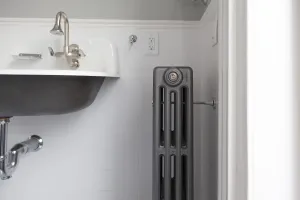 Plain Mercury 4 column cast iron radiator in a grey metallic finish with a wall stay, beside a wall-hung basin in a white-tiled bathroom.