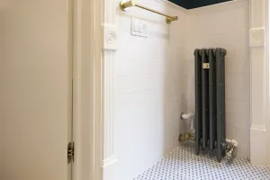 Narrow Mercury 4 column cast iron radiator in dark grey on feet, in a white-tiled bathroom alcove with a blue patterned floor and brass towel rail.
