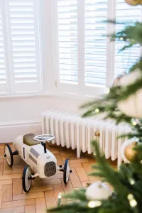 Low white Mercury 6 Column 360mm cast iron radiator with brass valve below shuttered bay window, toy car on parquet, Christmas tree in front.