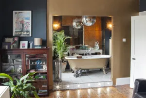 A gold roll-top bath on claw feet in a tiled alcove with antique mirror panels, palm and disco balls, beside a mahogany display cabinet.
