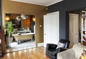 Living room with a gold-walled alcove holding a roll-top bath on claw feet, dark grey walls, white panelled doors and a black leather armchair.