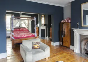 Dark blue-grey bedroom with a four-poster bed, grey armchair, walnut wardrobe and white marble fireplace on bare floorboards; no radiator visible.