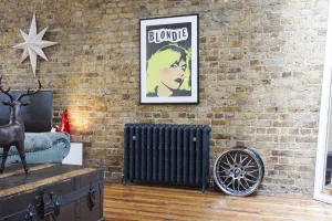 Ornate Rococo III cast iron radiator in Charcoal on feet against exposed brick, below a framed Blondie print, with a wheel and trunk on a wooden floor.