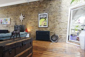 Ornate Rococo III 768mm cast iron radiator in Charcoal on feet against a bare brick wall, below a Blondie poster, on worn wooden floorboards.