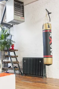 Ornate Rococo III cast iron radiator in Charcoal on feet against a white brick wall, beside a hanging punch bag on a wooden floor.