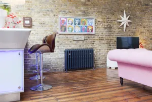 Ornate Rococo III cast iron radiator in Charcoal on feet against an exposed brick wall, with oak floorboards, bar stools and a pink sofa.