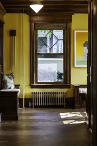 A plain Mercury 4 Column cast iron radiator in Bronze Gold under a wood-framed window, between oak window seats on a herringbone floor.