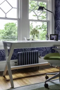 Plain Mercury 4 Column 460mm cast iron radiator in Caspian on feet, under a white desk by sash windows against navy floral wallpaper.