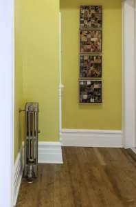 Narrow plain Mercury 4 Column cast iron radiator in Bronze Gold on feet with a valve and wall stay, in a yellow hallway with wooden floorboards.