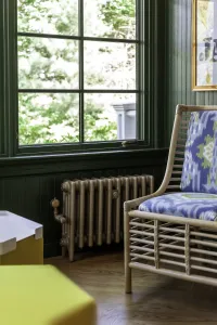 Plain Mercury 4 Column cast iron radiator in Bronze Gold on feet with a valve, under a dark green window beside a rattan armchair.