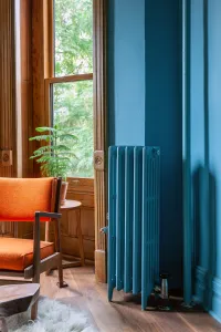 Tall, narrow Mercury 6 Column radiator on feet in Stone Blue, beside a wooden sash window, orange armchair and timber floor.