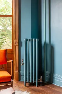 Narrow plain Mercury 6 Column cast iron radiator on feet in Stone Blue, matching the wall, beside an oak window frame and orange armchair.