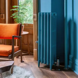 Plain Mercury 6 Column radiator in Stone Blue on feet with chrome valves, beside a blue wall, wooden window panelling and an orange armchair.