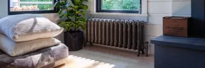 Ornate Rococo II 510mm cast iron radiator in Grey Bronze on feet beneath a window, with angled valve, plant and floor cushions nearby.