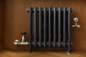 Mercury 3 column cast iron radiator in Natural Pewter on feet against a wood-panelled wall, with a nickel angled valve and thermostatic valve.