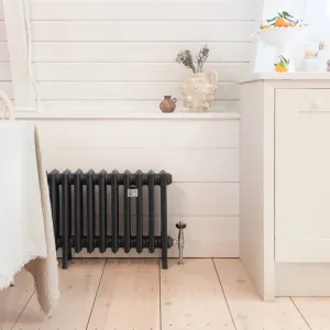 Grace 4 Column 500mm cast iron radiator in Farrow & Ball Black Blue No. 95 on feet, with a nickel valve, beside white panelling and pine boards.