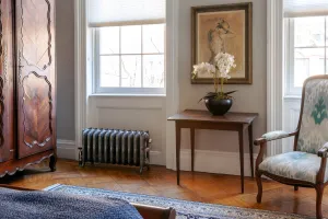Ornate Rococo III cast iron radiator in Etch-Coated Polish on feet beneath a sash window, beside a wooden side table on a parquet floor.