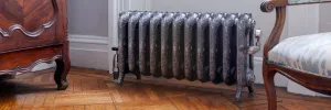 Ornate Rococo III cast iron radiator in Etch-Coated Polish on feet, with chrome valves, on a parquet floor between a carved cabinet and armchair.
