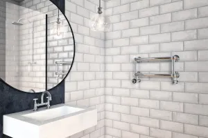 Small wall-mounted Elara towel rail with two chrome bars and valves on white brick tiles, beside a basin, round mirror and glass pendant light.