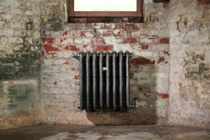 Ornate Rococo III 768mm cast iron radiator in Full Polish, wall-hung below a window on a peeling red brick wall, with brass valves.