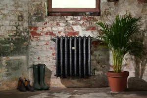 Ornate Rococo III cast iron radiator, wall-hung below a window on worn brick, with valves, wellington boots and a potted palm beside it.