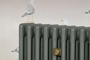 Close-up of a plain grey Mercury 4 Column cast iron radiator with a brass bee-motif Whitworth stay, against seagull-print wallpaper.