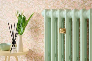 Plain Mercury cast iron radiator in Little Greene Pea Green 91 with a brass Whitworth wall stay, against pink floral wallpaper beside a side table.