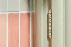 Close-up of an Emmeline cast iron radiator column in Pebble Grey, with the raised EMMELINE casting, beside pink and cream wall tiles.