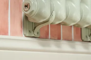Close-up of an Emmeline I cast iron radiator in Pebble Grey resting on matching wall brackets above a cream skirting board, pink tiled wall behind.