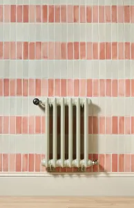 Emmeline I 665mm cast iron radiator in Pebble Grey, wall-hung on stays, with Windsor thermostatic valve against pink and cream striped tiles.