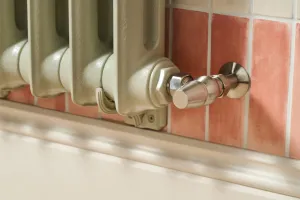 Close-up of the end of an Emmeline I cast iron radiator in Pebble Grey with a satin nickel angled valve against terracotta wall tiles.