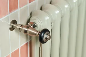 Close-up of an Emmeline I cast iron column radiator in Pebble Grey with an angled brushed metal valve, against pink and cream wall tiles.