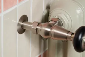 Close-up of a brushed nickel angled valve with black knob on a Pebble Grey Emmeline cast iron radiator against pink and cream wall tiles.