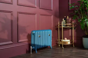 Emmeline II 470mm cast iron radiator on feet in a blue finish with a silver valve, against a burgundy panelled wall beside a brass drinks trolley.