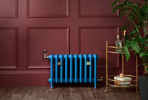 Bright blue Emmeline II 470mm cast iron radiator on feet with brass valves, against a burgundy panelled wall beside a brass drinks trolley.