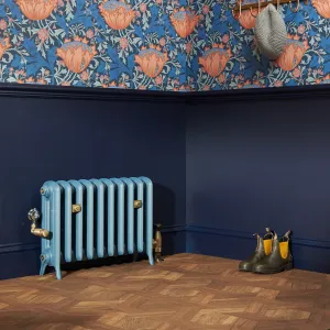 Light blue Emmeline II cast iron radiator on feet with brass valves, against a navy wall below floral wallpaper, on a parquet floor.