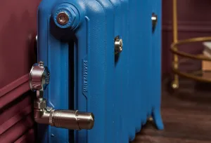 Close-up of a blue Emmeline II cast iron radiator on feet with a polished nickel thermostatic valve, against a burgundy panelled wall.