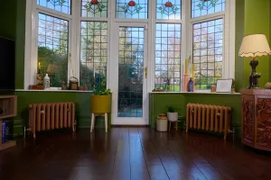 Two low Emmeline III cast iron radiators in Little Greene Affogato 342 on feet beneath a stained glass bay window in a green-walled room with dark floorboards.