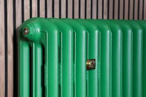 Close-up of an Emmeline III cast iron radiator in Farrow & Ball Danish Lawn green with a brass wall stay, against slatted timber panelling.