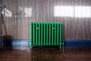 Emmeline III cast iron radiator in Farrow & Ball Danish Lawn green on feet, with brass valves, against slatted wood panelling on a dark floor.