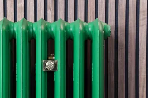 Close-up of an Emmeline III cast iron radiator in Farrow & Ball Danish Lawn green with a brass wall stay, against dark wooden slat panelling.