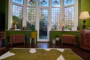 Two low Emmeline III cast iron radiators in Little Greene Affogato 342 under a bay window with stained glass, green walls and a dark wood floor.