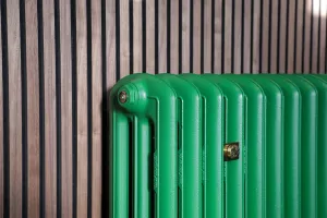 Close-up of an Emmeline III cast iron radiator in Farrow & Ball Danish Lawn green with a brass wall stay, against a slatted walnut wall.