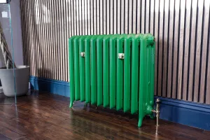 Green Emmeline III cast iron radiator on feet with brass wall stays and valve, against slatted wood panelling above a blue skirting and dark wood floor.